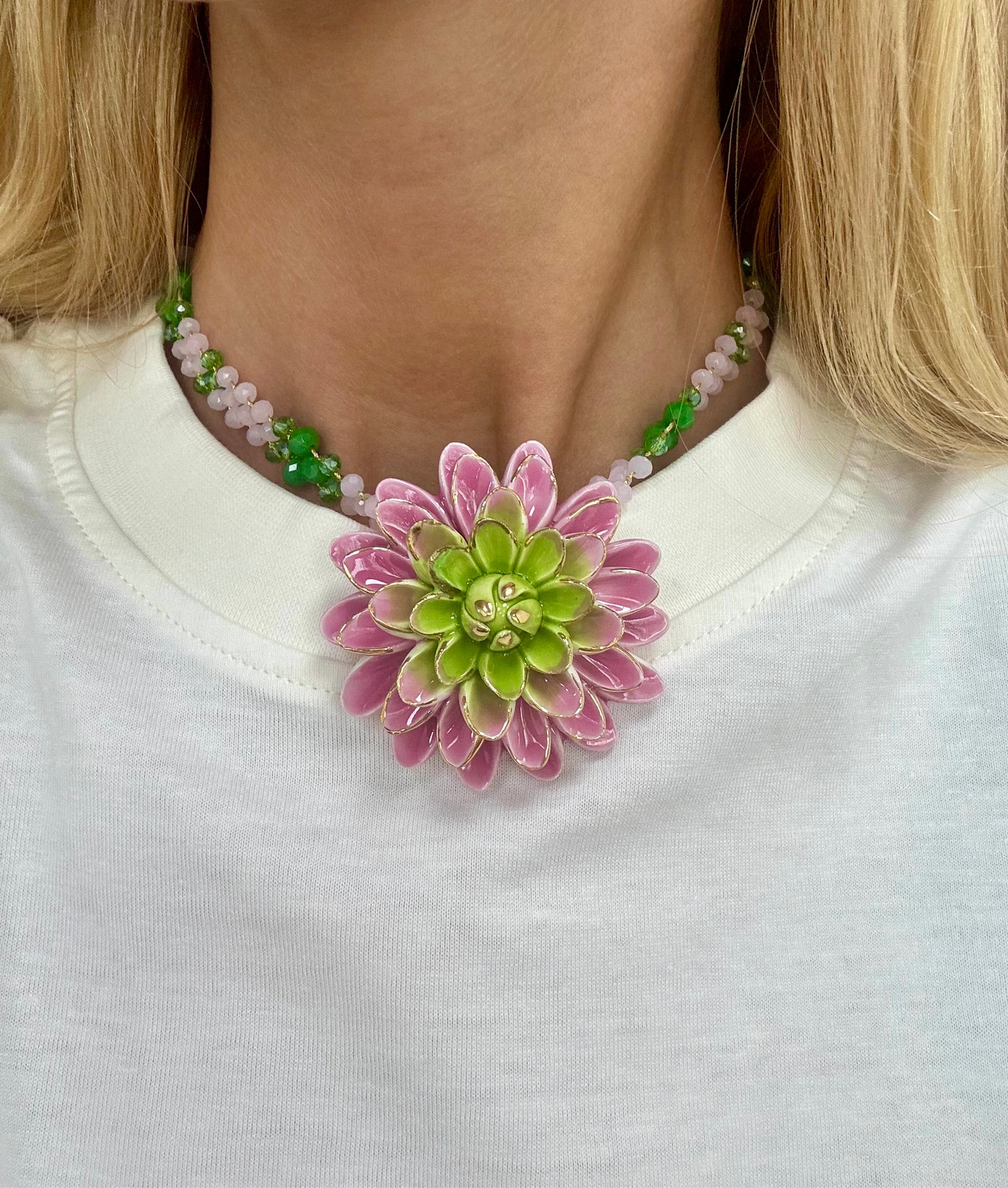 Collar con cuentas verdes y rosas pastel, con un gran colgante de flor tipo dalia esmaltada en tonos rosados y verde lima, sobre fondo blanco. Combinado con camiseta blanca básica. Cashfana.