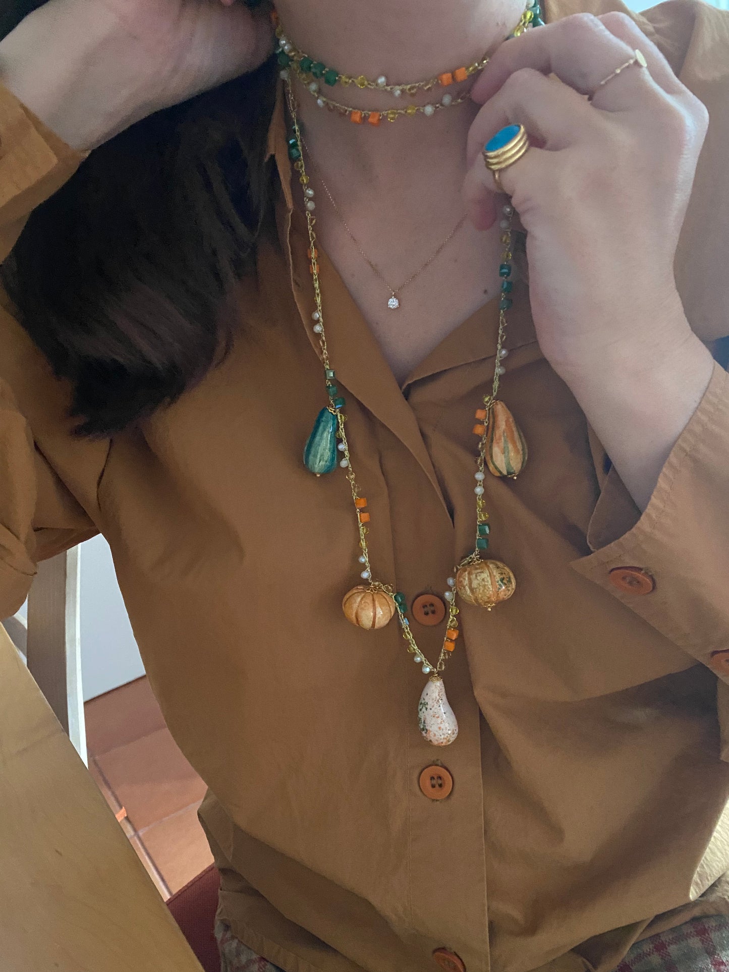 Collar largo con cuentas en tonos naranja y verde, charms de cerámica en forma de calabazas, con doble vuelta al cuello con camisa marrón.
Cashfana.