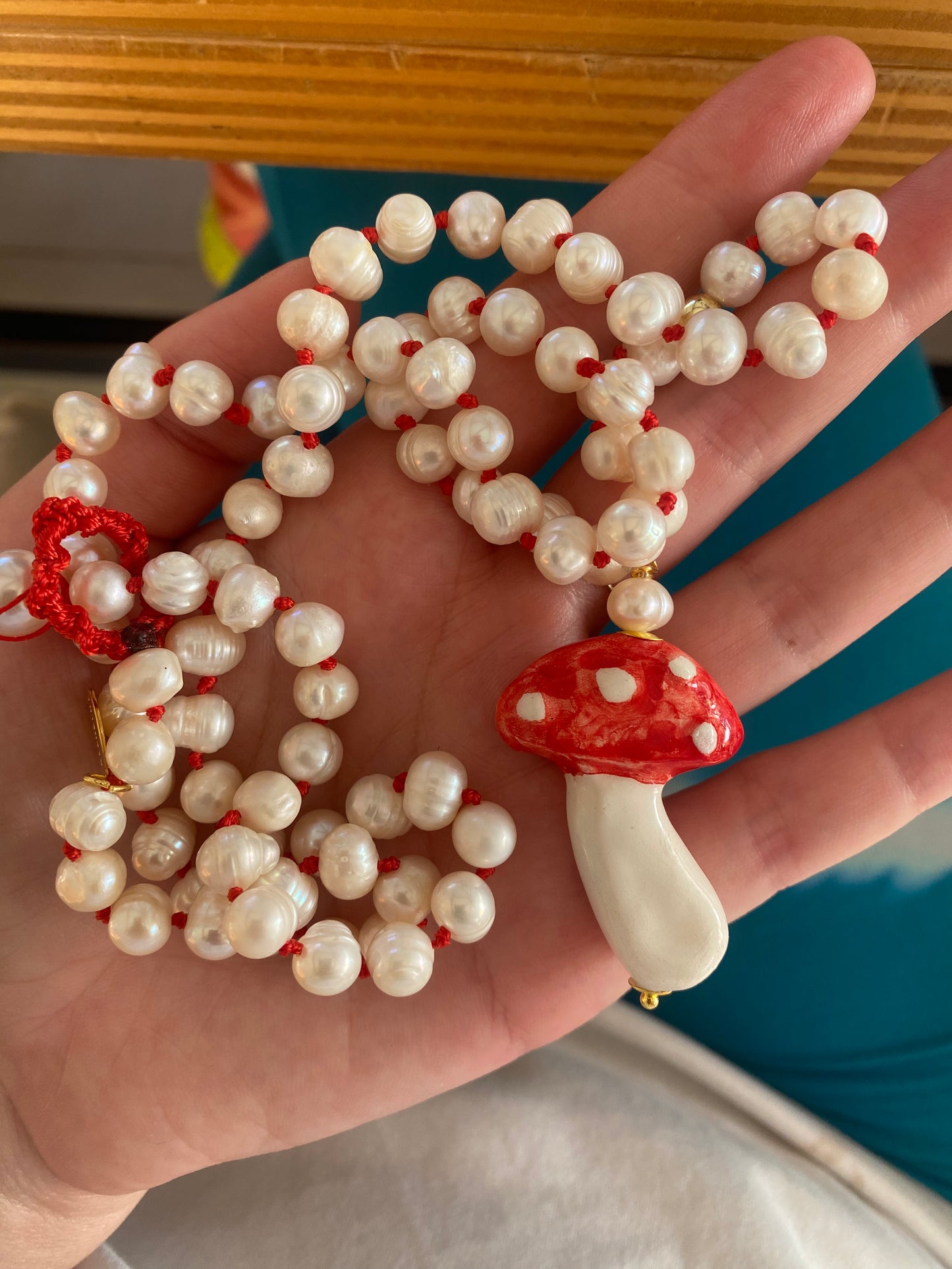 Collar artesanal de perlas naturales y nudos rojos intercalados con colgante esmaltado en forma de hongo rojo y blanco. Sobre una mano. Cashfana.