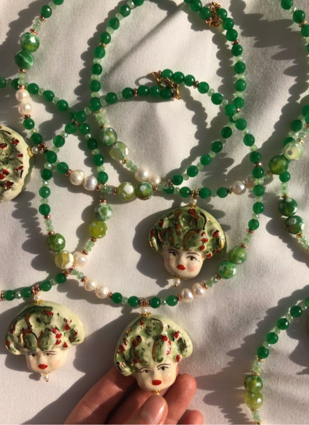 Collar corto con cuentas verdes, perlas y colgante de cerámica pintado a mano con una carita adornada de hojas y uvas rojas. Cierre dorado con extensión. Sobre fondo blanco con muchos collares.