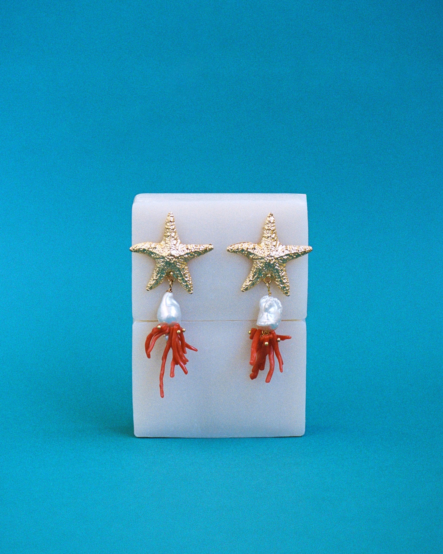 Pendientes dorados en forma de estrella de mar con colgantes de perla y coral rojo, diseño veraniego inspirado en el mar Mediterráneo. Sobre esponja blanca.