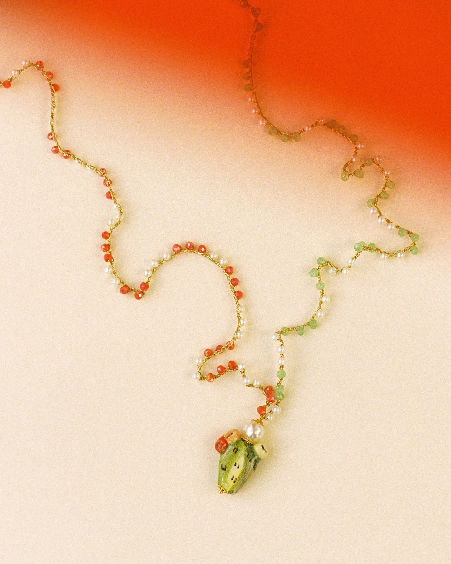 Collar largo con cuentas verdes y coral y colgante de cactus de cerámica esmaltada en verde. Pieza artesanal de Cashfana. Sobre fondo rojo y blanco.