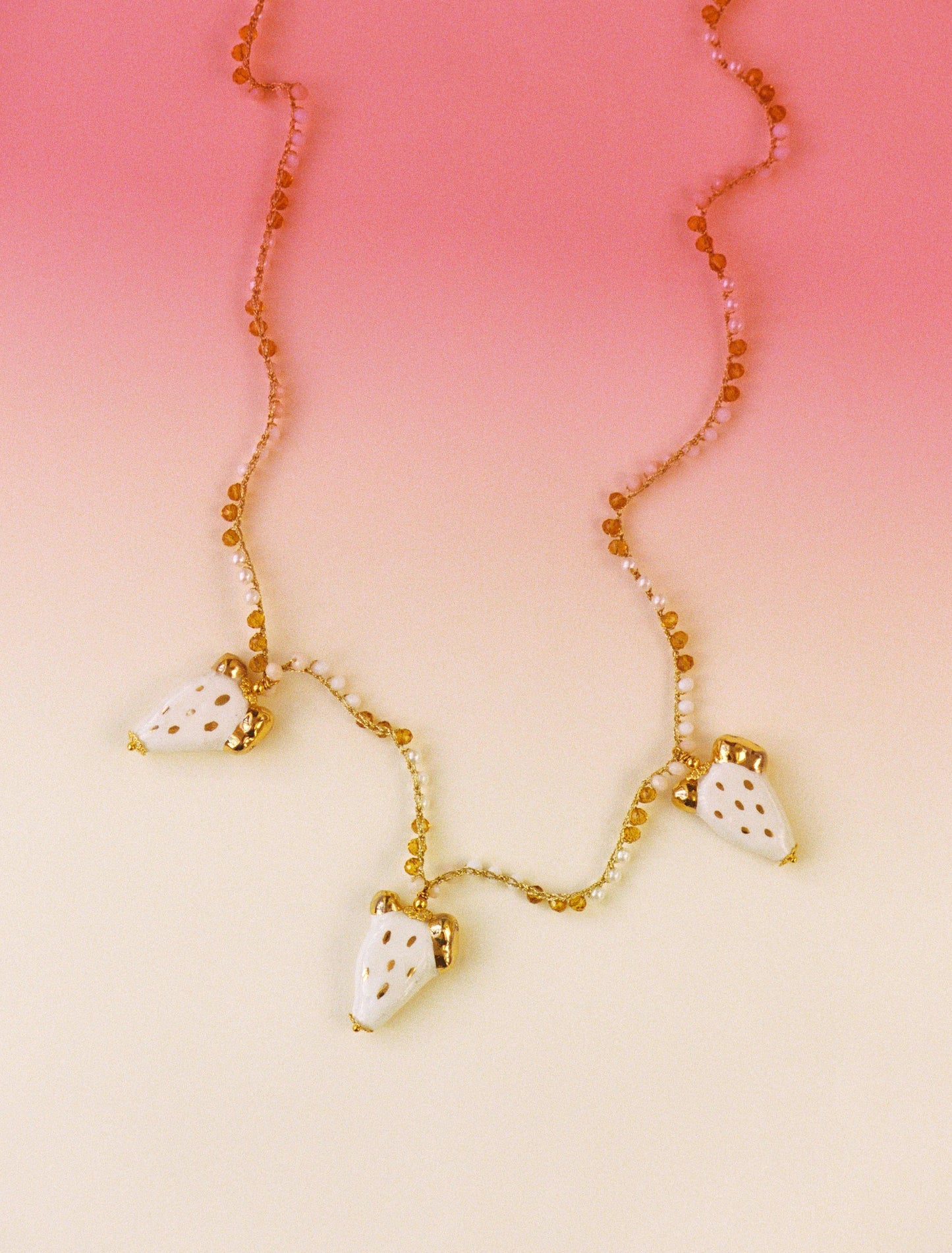 Collar largo con cuentas doradas y blancas, y tres colgantes de cactus de cerámica blanca pintados a mano. Diseño artesanal de Cashfana. Sobre fondo blanco y rosa.