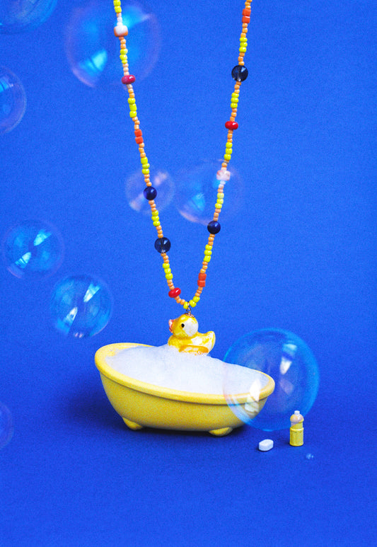 Collar largo con cuentas amarillas, naranjas, azules y rojas, y charm de patito de goma amarillo. Fondo azul con burbujas y una bañera.