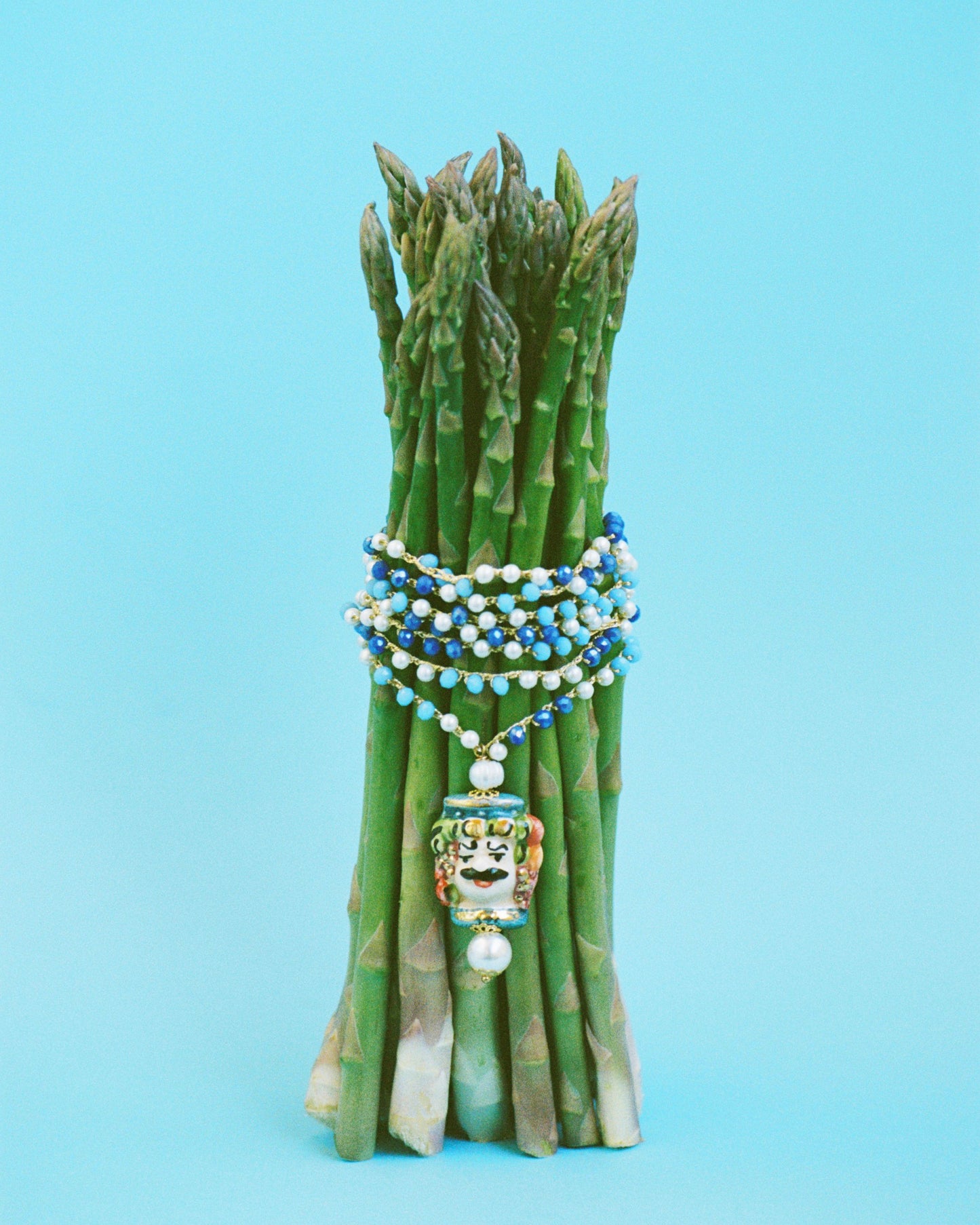 Collar largo con cuentas en azul y blanco, cierre dorado con perla y colgante de cerámica esmaltada con rostro masculino pintado a mano. Cashfana. Sobre fondo azul con esparragos.