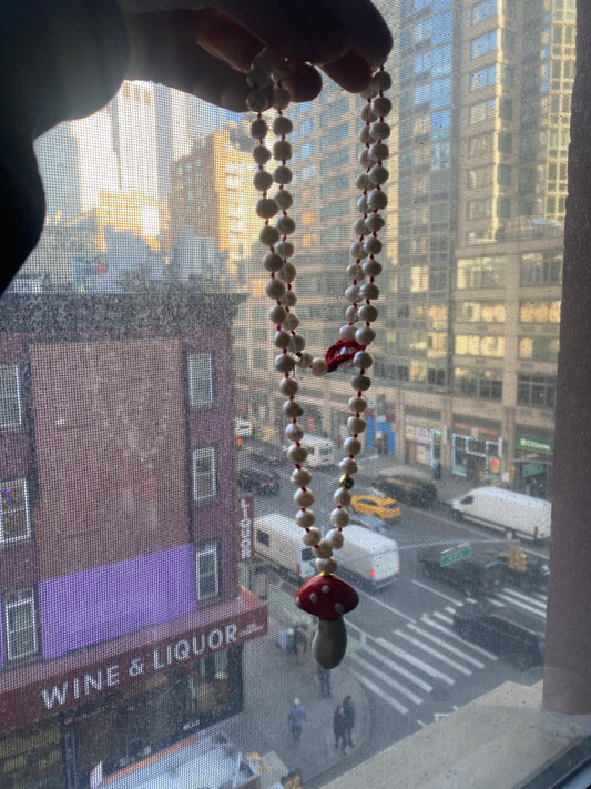 Collar artesanal de perlas naturales y nudos rojos intercalados con colgante esmaltado en forma de hongo rojo y blanco. Fondo ciudad. Cashfana.