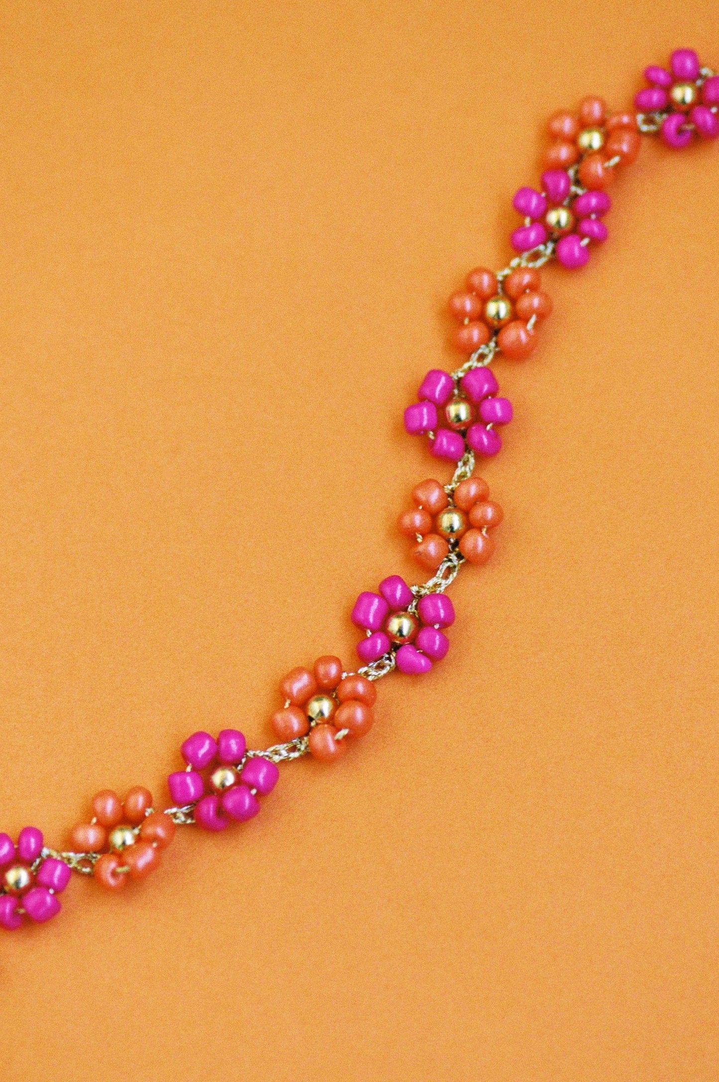 Choker Nardo de flores hechas a mano con cuentas fucsia brillante y naranja mandarina, con detalles dorados, sobre fondo naranja. Cashfana.