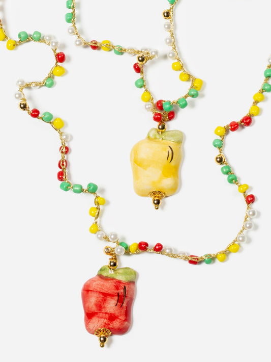 Collar largo con cuentas multicolores y dos colgantes esmaltados de manzana roja y manzana amarilla, sobre fondo blanco. Cashfana.