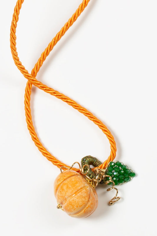 Collar de hilo trenzado naranja con perlas en los extremos y colgante de cerámica en forma de nuez con hoja verde, y cierre con perlas, sobre fondo blanco. Cashfana.