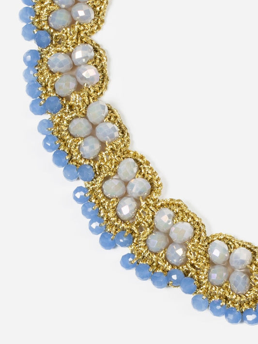 Collar tipo gargantilla bordada a mano en hilo dorado con cuentas grises y cristales azul celeste, formando un diseño simétrico sobre fondo blanco. Cashfana.