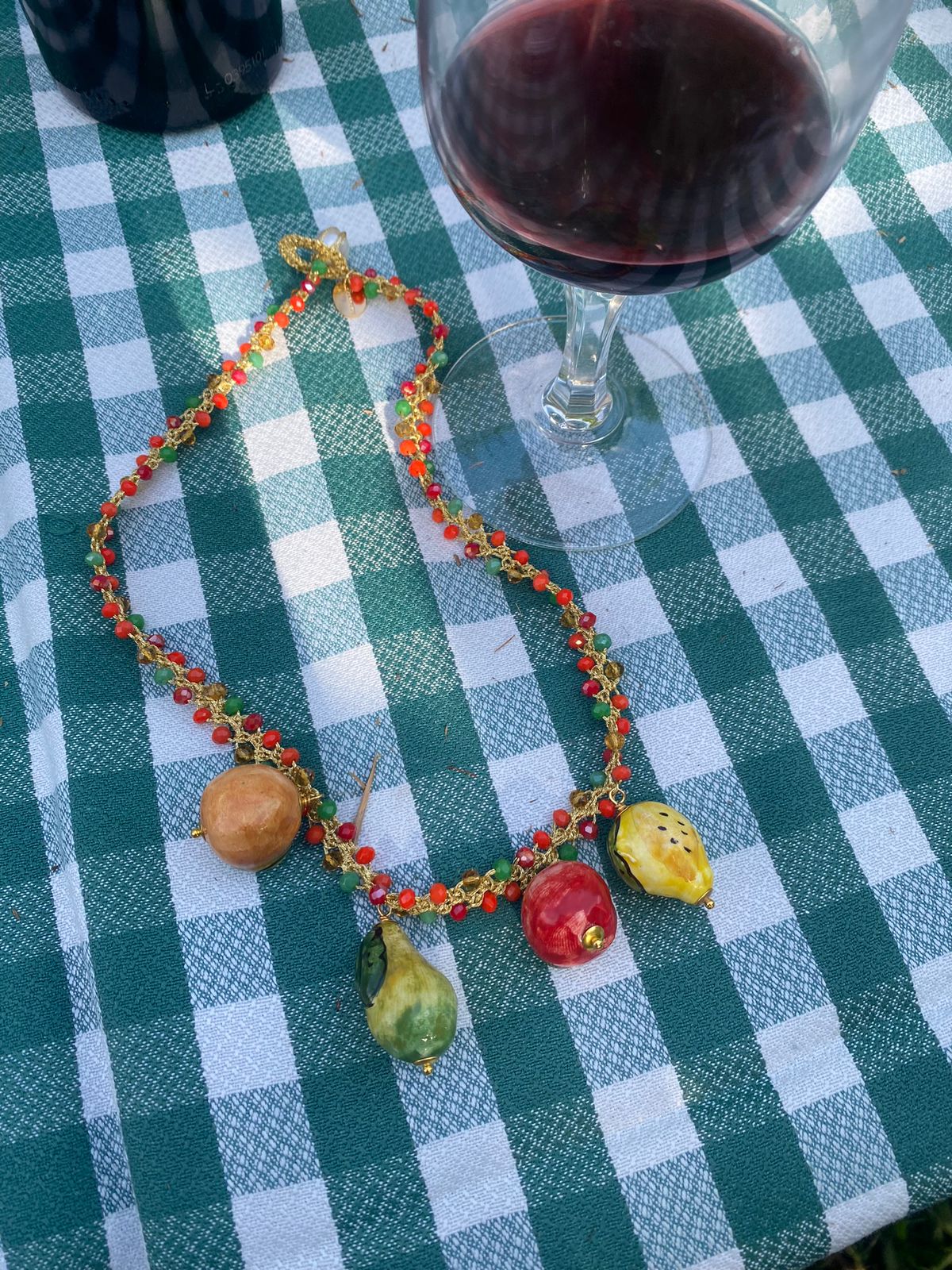 Collar de cuentas multicolor con charms colgantes de frutas esmaltadas: manzana roja, naranja, limón y pera, sobre mantel a cuadros. Cashfana.