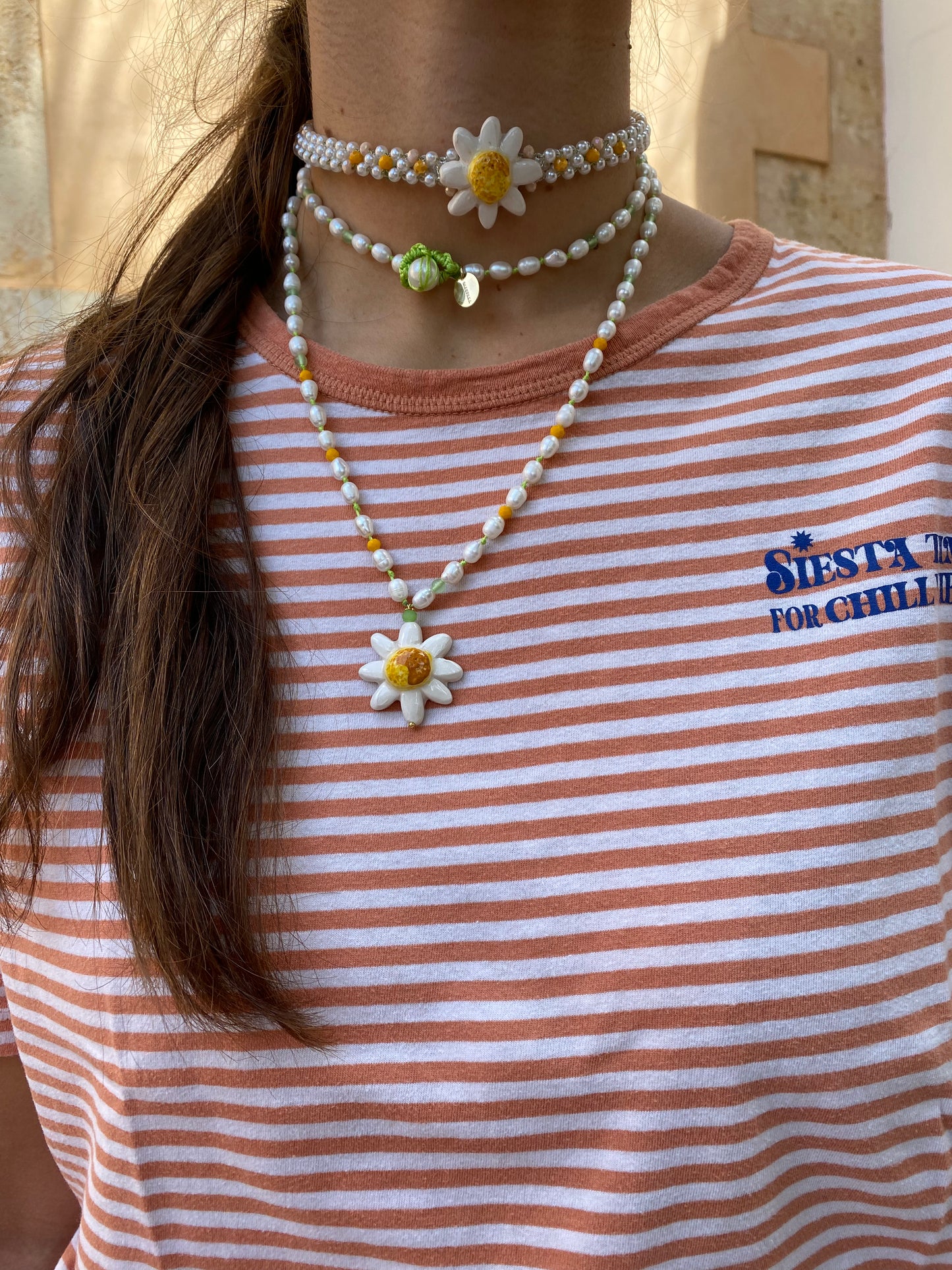 Collar largo con perlas, cuentas en tonos lavanda, verde menta y amarillo pastel, y colgante de flor esmaltada en blanco con centro dorado, sobre camiseta de rayas con la gargantilla Margherita. Cashfana.