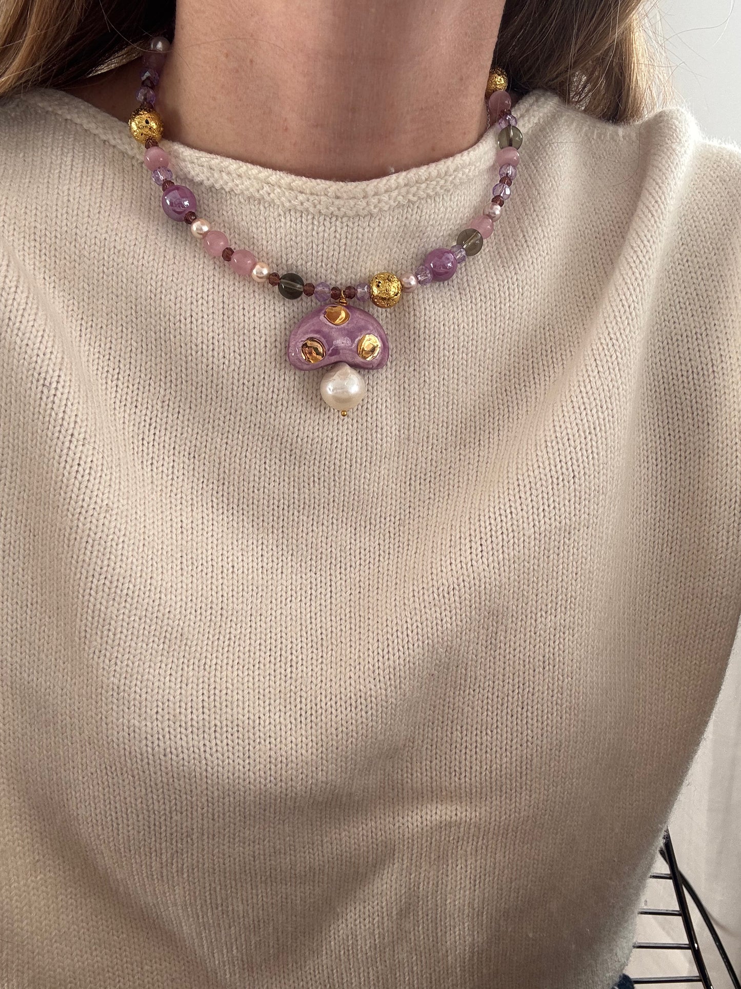 Collar corto con cuentas lila, rosadas y doradas, y pieza de cerámica en forma de hongo violeta con perlita, combinado con jersey beige. Cashfana.