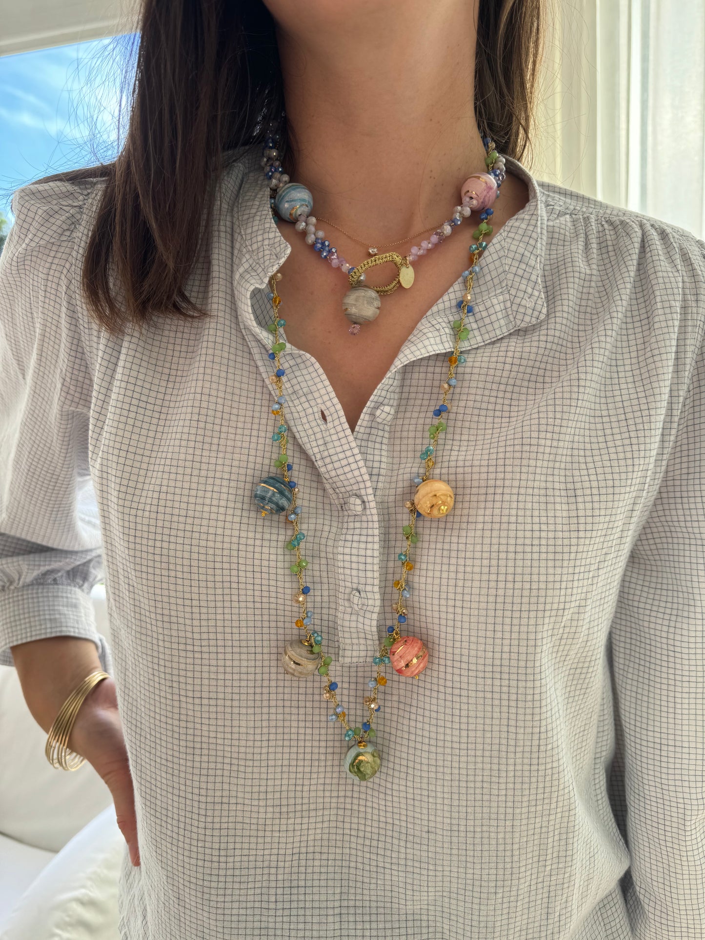 Collar largo con cuentas en tonos lavanda, turquesa, verde lima, mostaza y blanco perlado, decorado con cinco esferas de cerámica pintadas a mano en azul, rosa, gris, marrón y amarillo. Cashfana.
Con una vuelta más suelta sobre el cuello y otra más larga que luce los planetas.