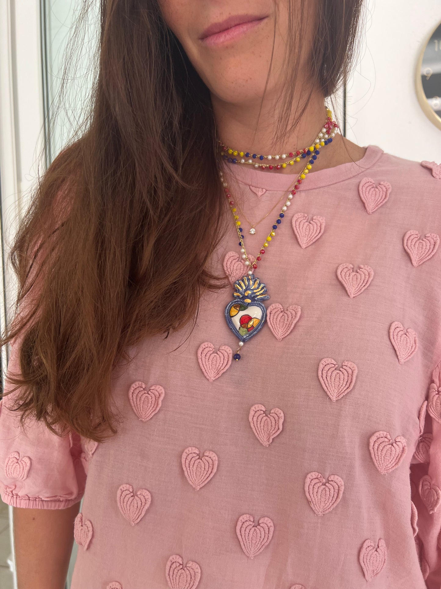 Collar largo con dos vueltas al cuello con cuentas multicolores y colgante central de corazón azul en cerámica esmaltada con flama pintada a mano y detalles dorados, sobre camiseta rosa con estampado de corazones. Cashfana.