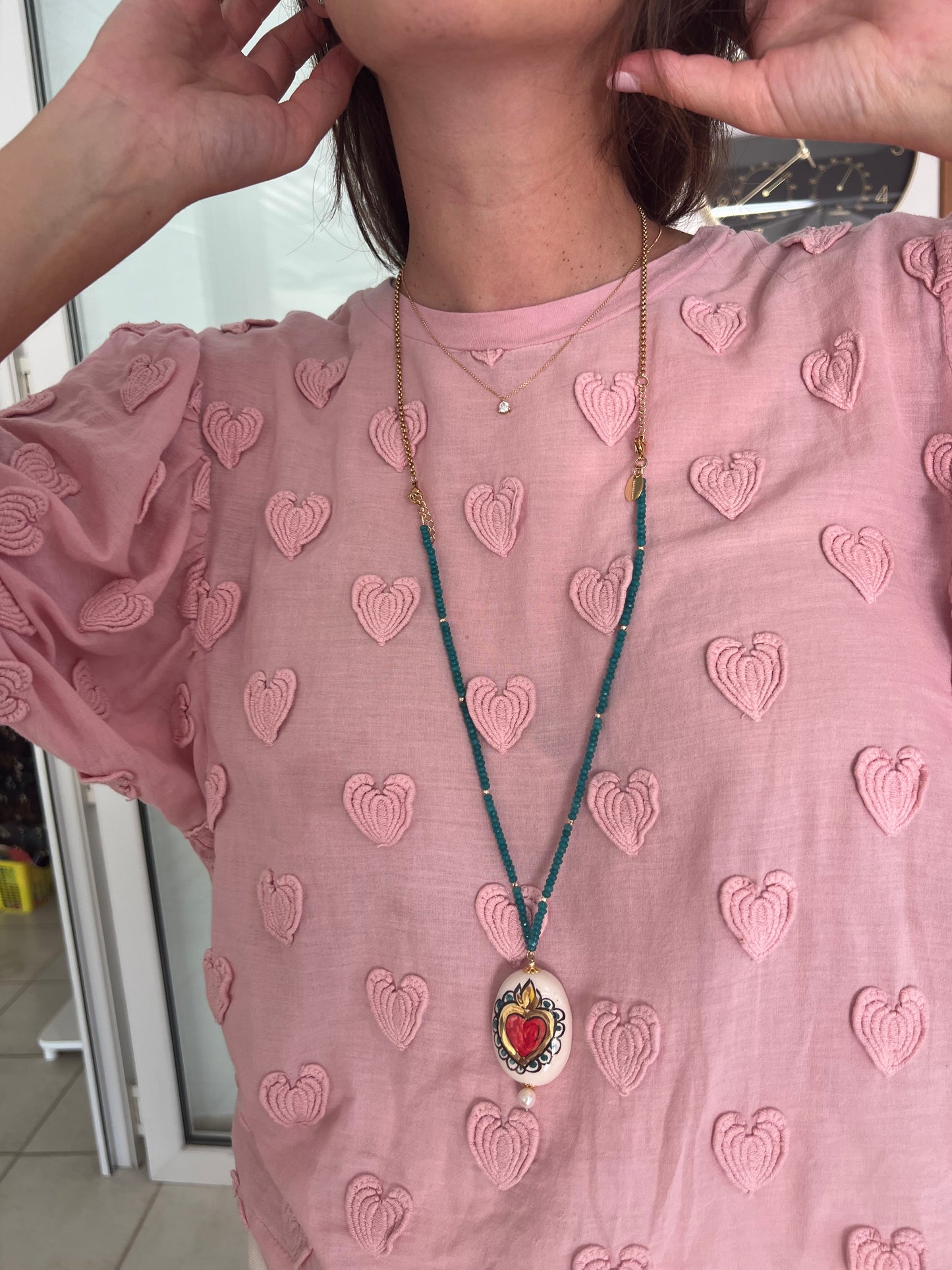 Collar Cuore Biancho largo con cuentas azul turquesa y corazón sobre fondo blanco en pieza de cerámica. Sobre camiseta rosa con estampado de corazones.