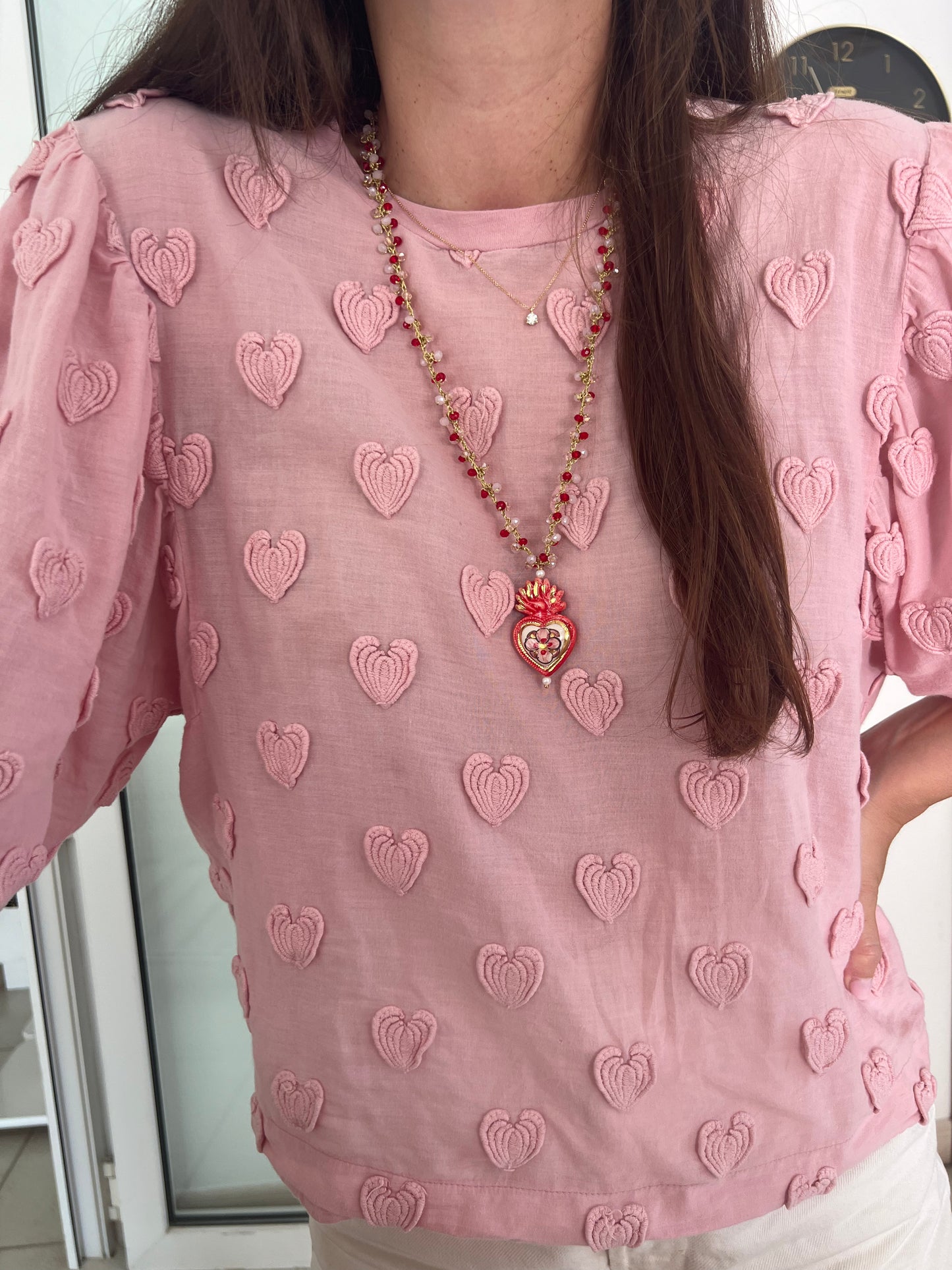 Collar de largo medio con cuentas rojas y doradas y colgante de cerámica en forma de corazón rojo decorado con flores y corona de fuego, sobre fondo blanco. Sobre camiseta rosa con estampado de corazones. Cashfana.