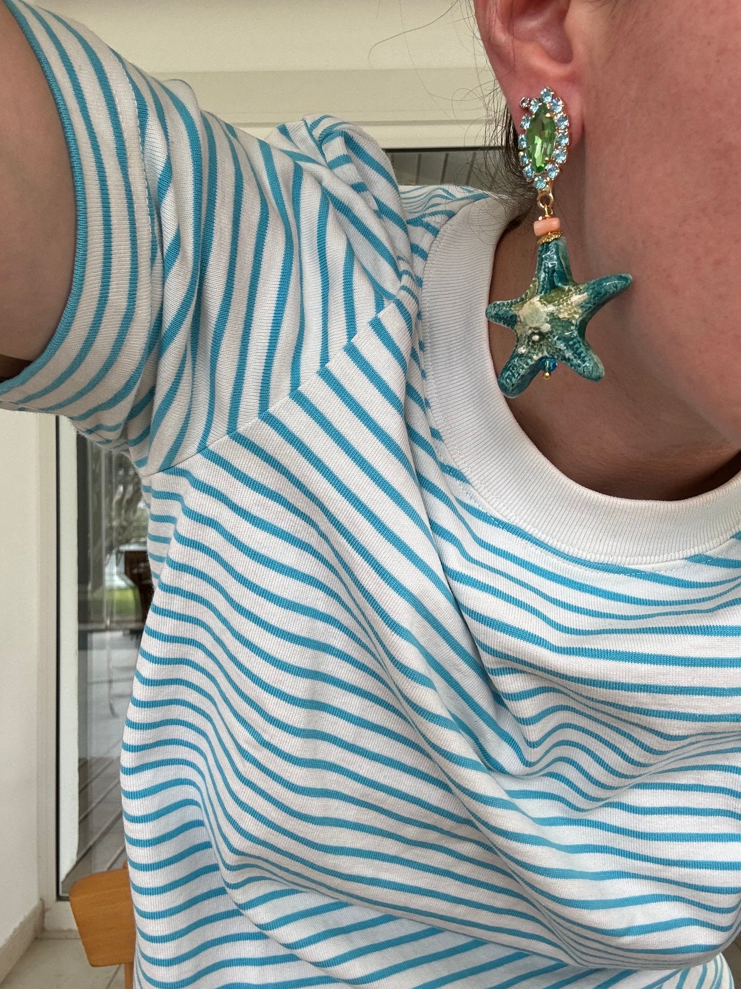 Pendientes con forma de estrella de mar en tonos verdes y turquesa de cerámica, colgantes de una base de cristal verde rodeado de piedras azul zafiro.
Combinado con una camiseta azul y blanca a rayas.