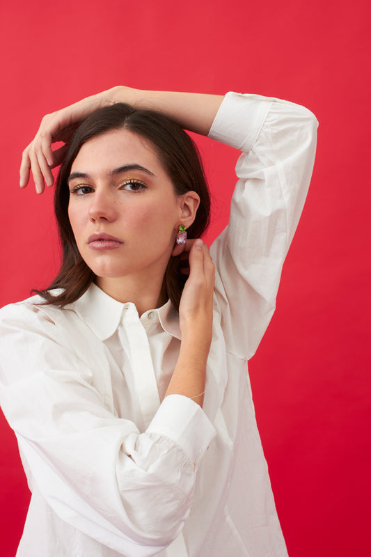 Pendientes pequeños con cristales rectangulares en rosa y dorado, combinados con piedras redondas en azul y verde. Sobre fondo rojo con modelo con camisa blanca.
