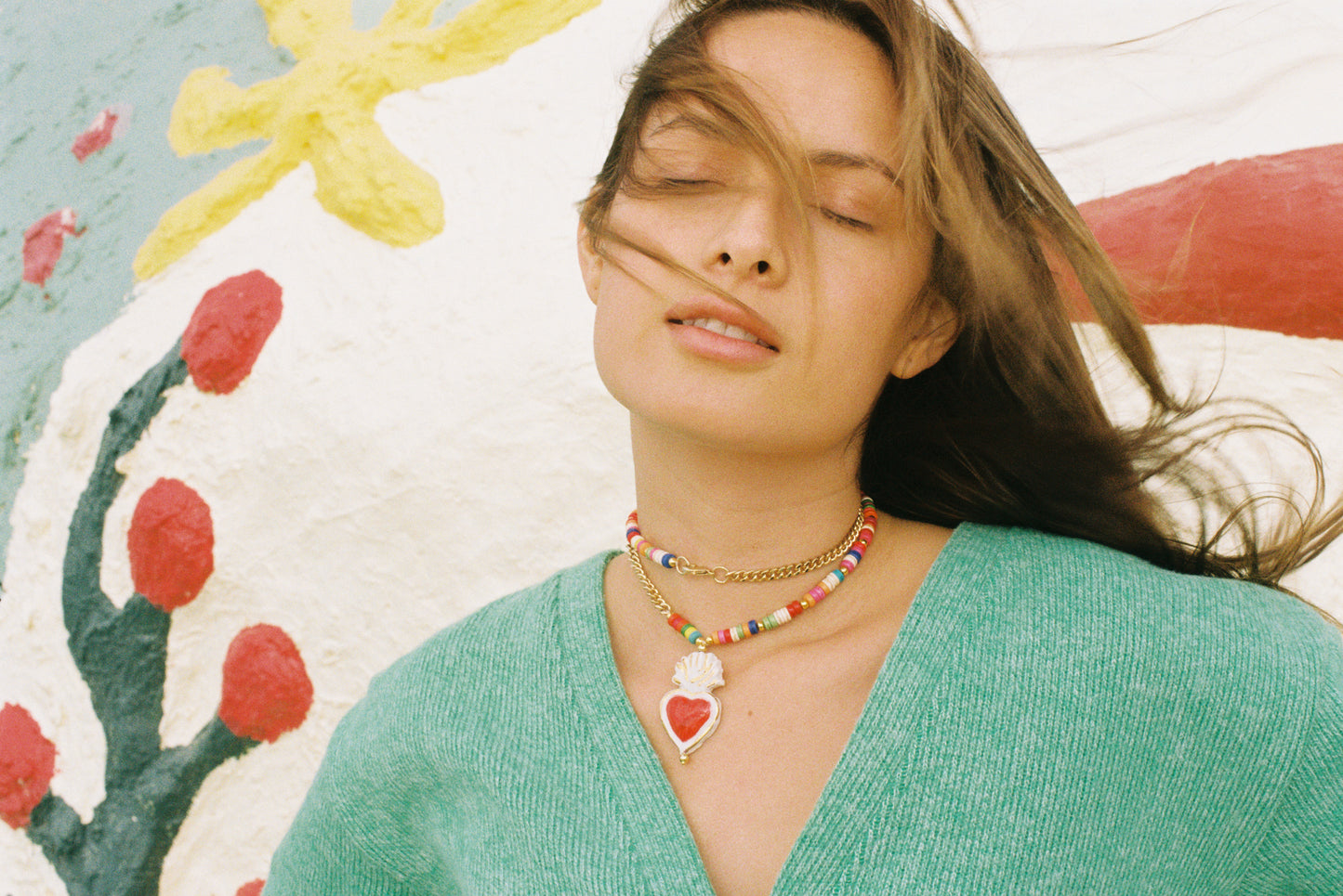 Modelo con jersey verde, pelo volado al viento y collar de cerámica Cuore Landia, con colgante de cerámica en forma de corazón rojo coronado por llama, sobre fondo blanco.
Cashfana.