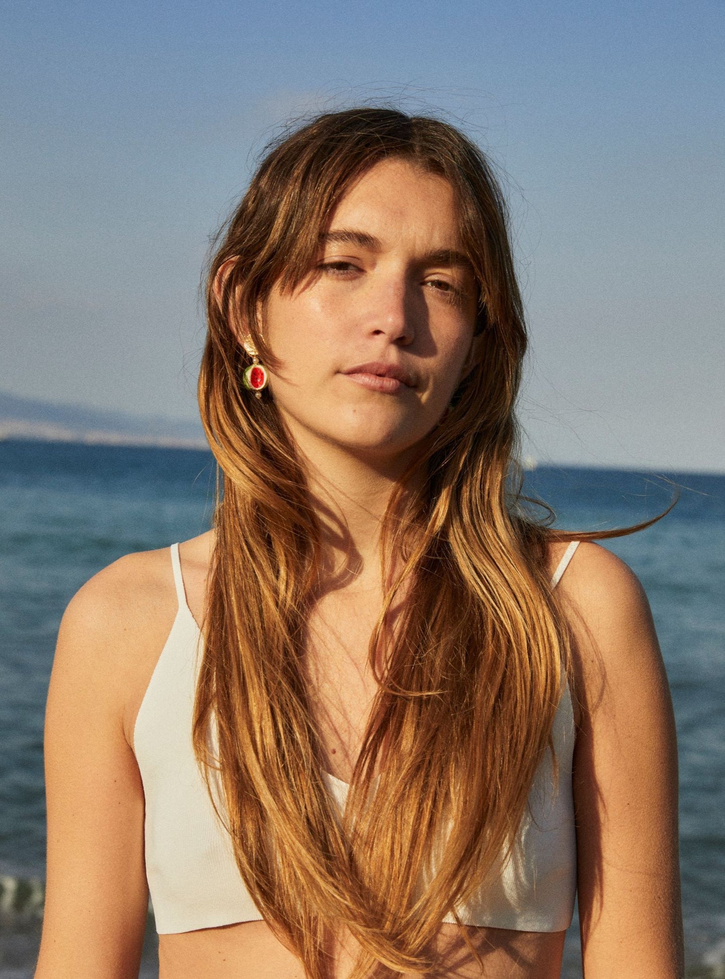 Pendientes con disco dorado y colgante de cerámica con diseño de sandía pintada a mano sobre fondo verde. En la playa con bikini blanco.