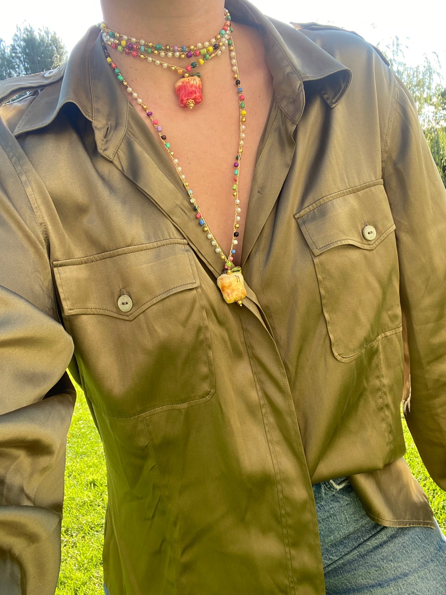 Collar largo con cuentas multicolores y dos colgantes esmaltados de manzana roja y manzana amarilla, sobre cuelllo con blusa verde satinada. Cashfana.