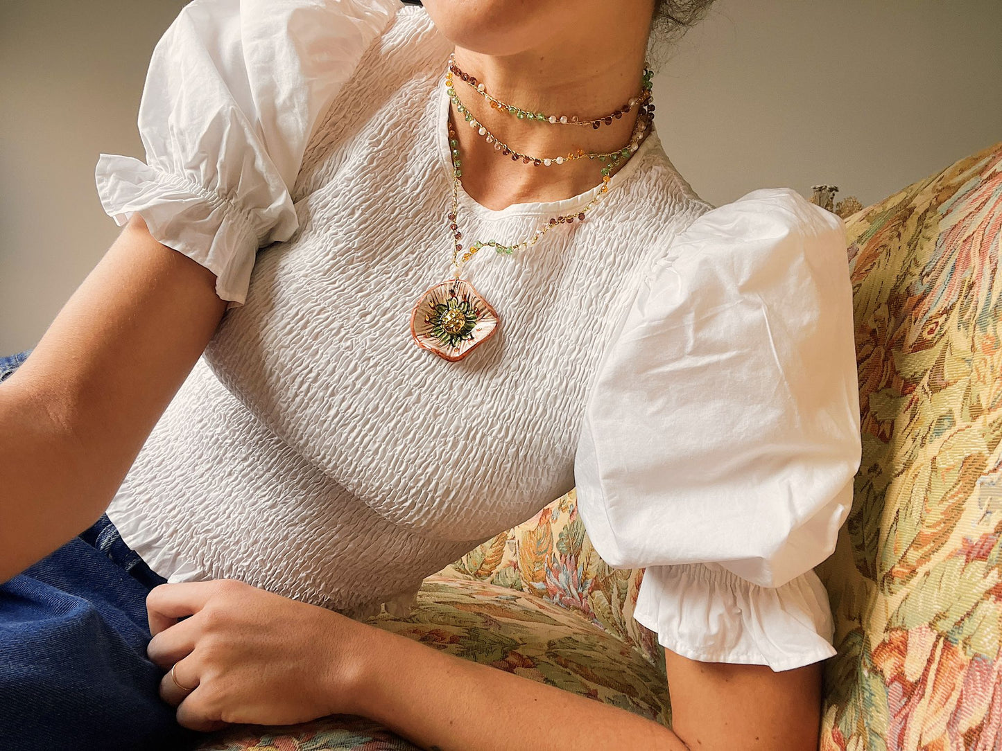 Collar Miranda largo con colgante esmaltado en forma de sol en tonos dorado, verde y terracota, cadena de cuentas en verde, lila, blanco y amarillo, sobre blusa blanca. Cashfana.