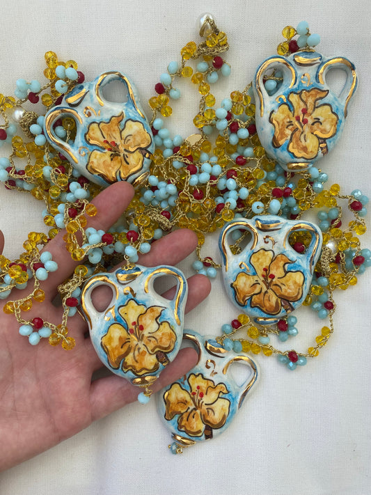 Varios collares Vase largos con colgante en forma de jarrón esmaltado con flor central en tonos crema y coral, cadena de flores miniatura en azul cielo, rojo y amarillo mostaza, sobre fondo blanco. Cashfana.
