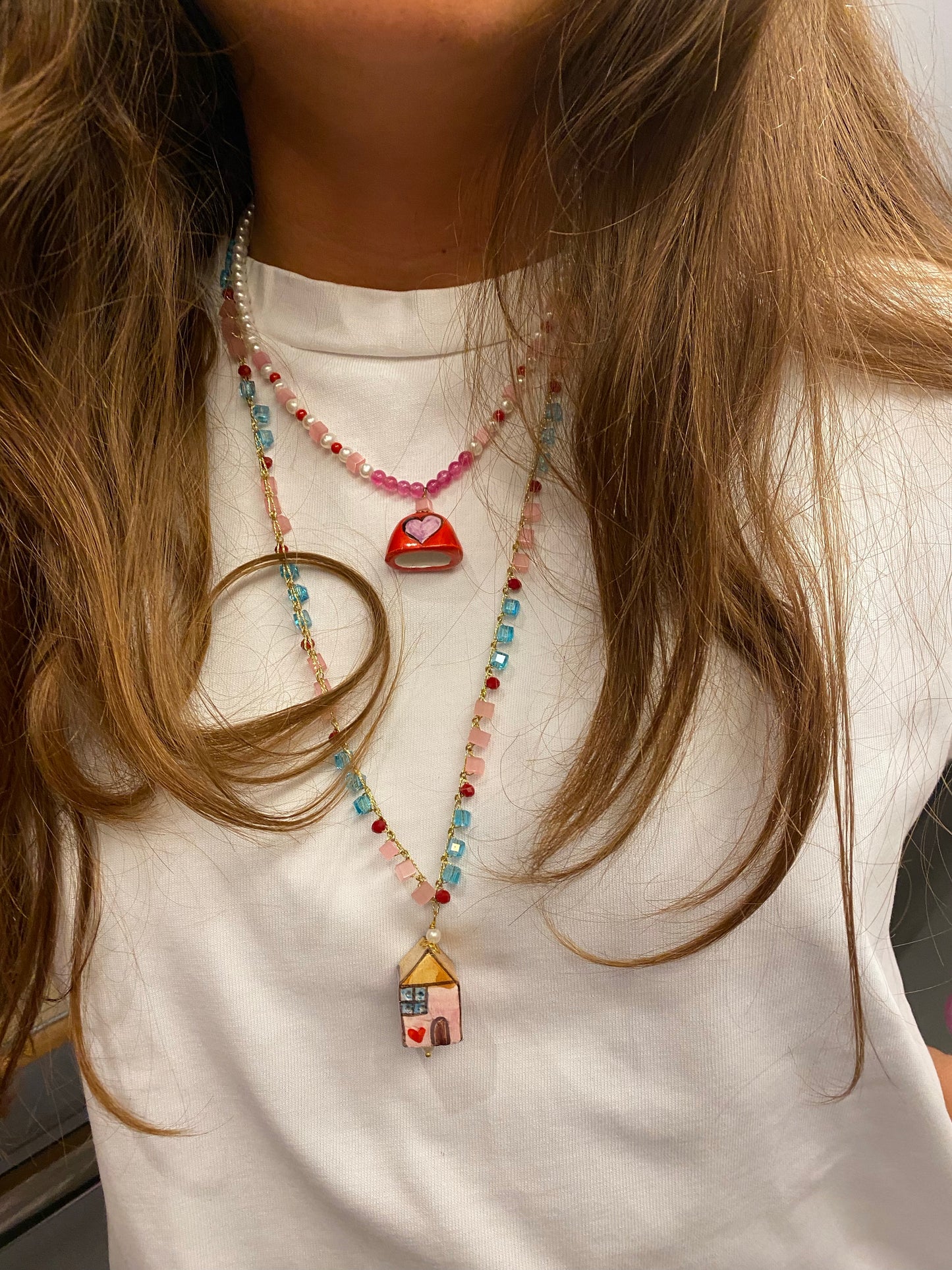 Collar corto de cuentas blancas, rosas y rojas con colgante de cerámica en forma de campana roja decorada con corazón rosa, sobre fondo blanco. Combinado con el collar La Casita sobre camiseta blanca. Cashfana.