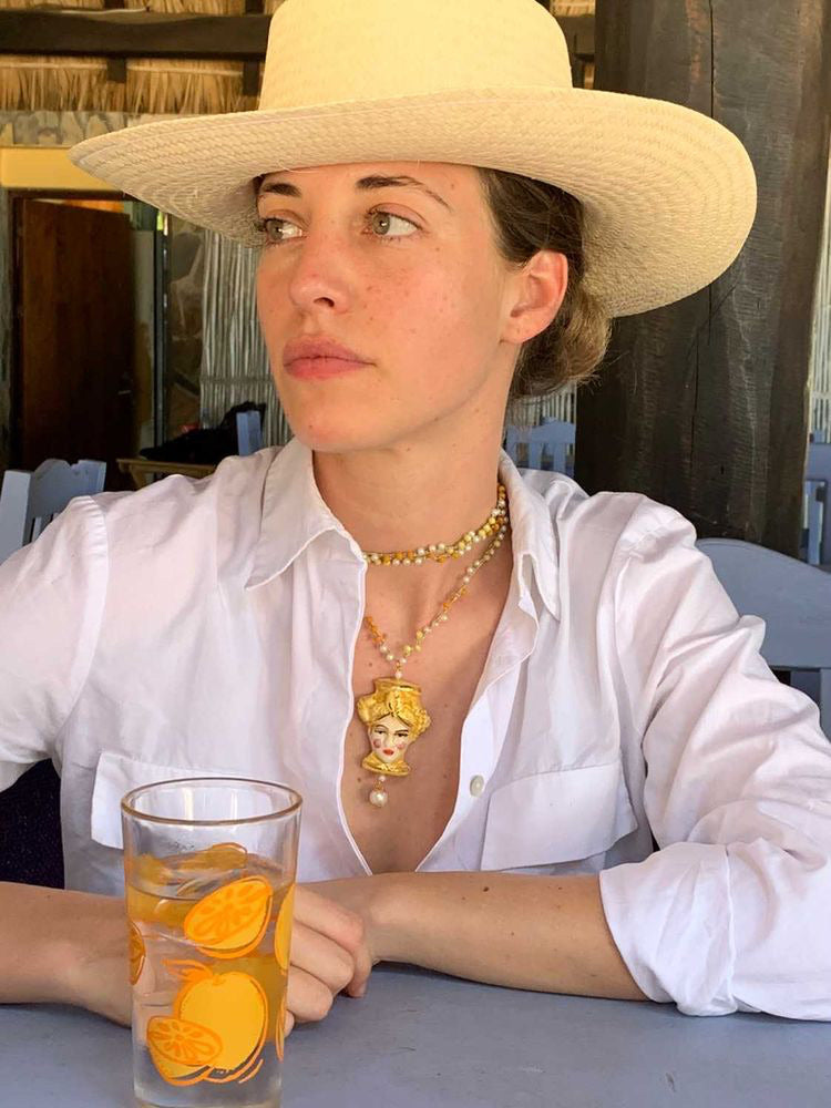 Collar largo con cuentas color miel y perlas naturales, con colgante de cerámica pintado a mano con rostro femenino y detalles dorados. Cashfana. Combinada con camisa blanca abierta.