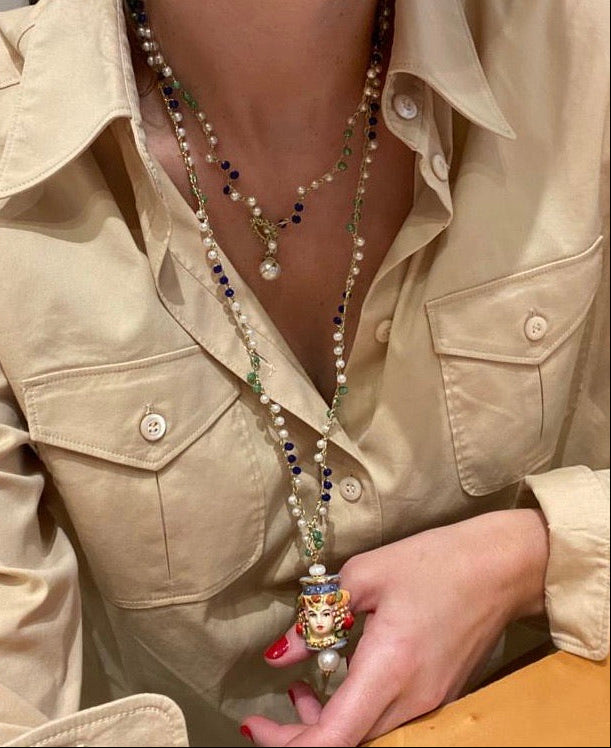 Collar largo con cuentas verdes, azul marino y perlas pequeñas, cierre dorado con perla y colgante de cerámica de rostro femenino pintado a mano. Con doble vuelta al cuello y camisa beige abierta.