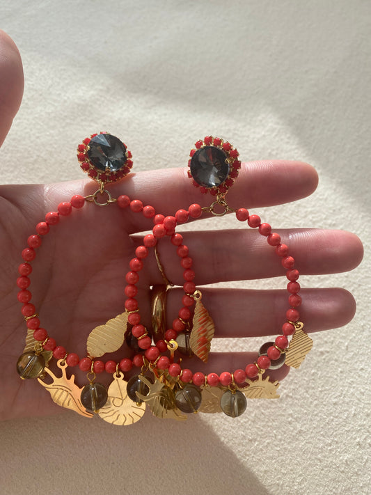Pendientes de aro coralino con colgantes dorados marinos y piedra negra central rodeada de cuentas rojas.