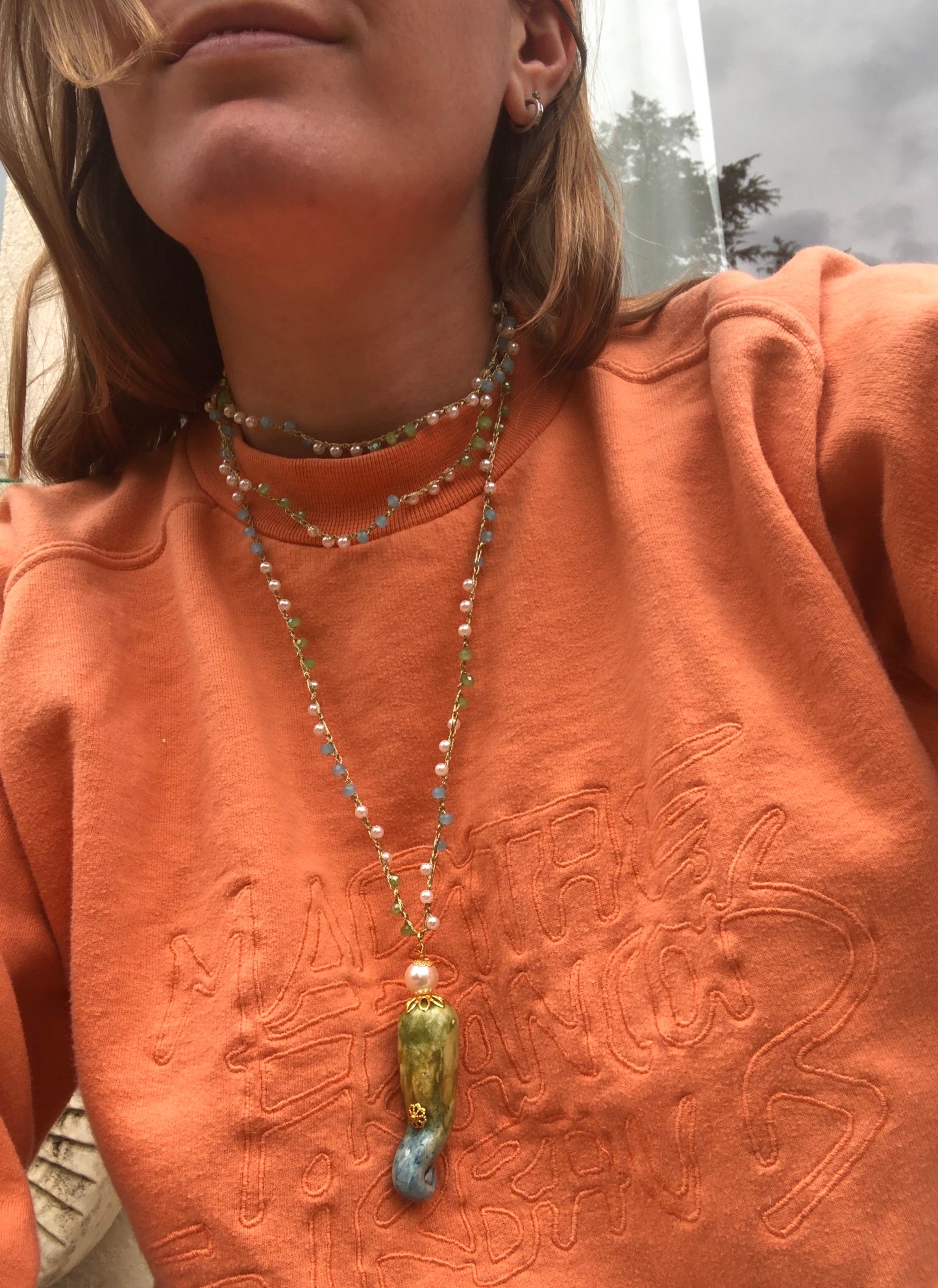 Collar largo con cuentas finas y colgante esmaltado en forma de peperone verde, amarillo y azul, combinado con sudadera naranja y dos vueltas al cuello. Cashfana.