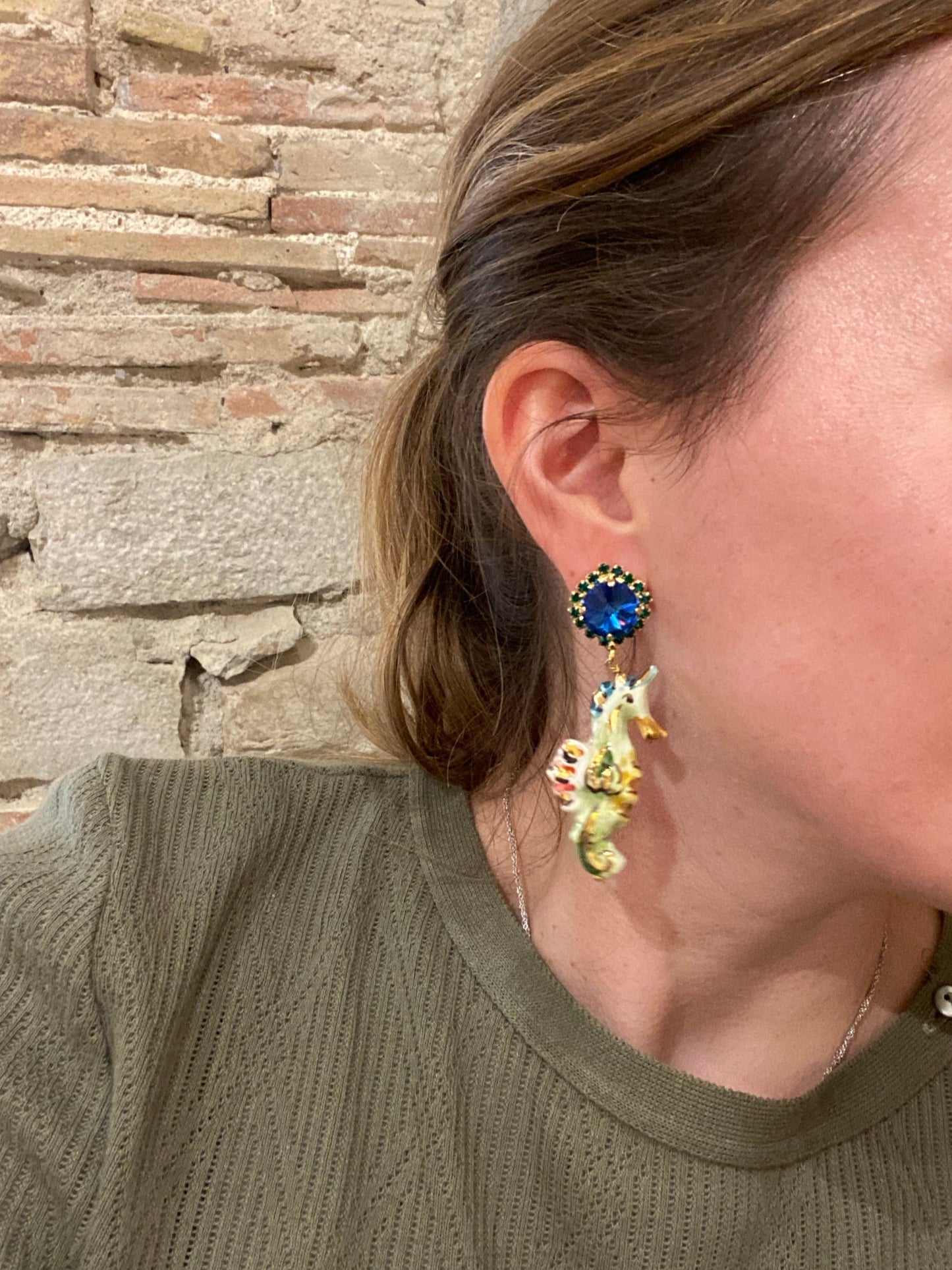 Pendientes caballito de mar esmaltado con base de cristal azul y detalles pintados a mano en tonos marinos, verde, coral, dorado y naranja. Con camiseta verde.