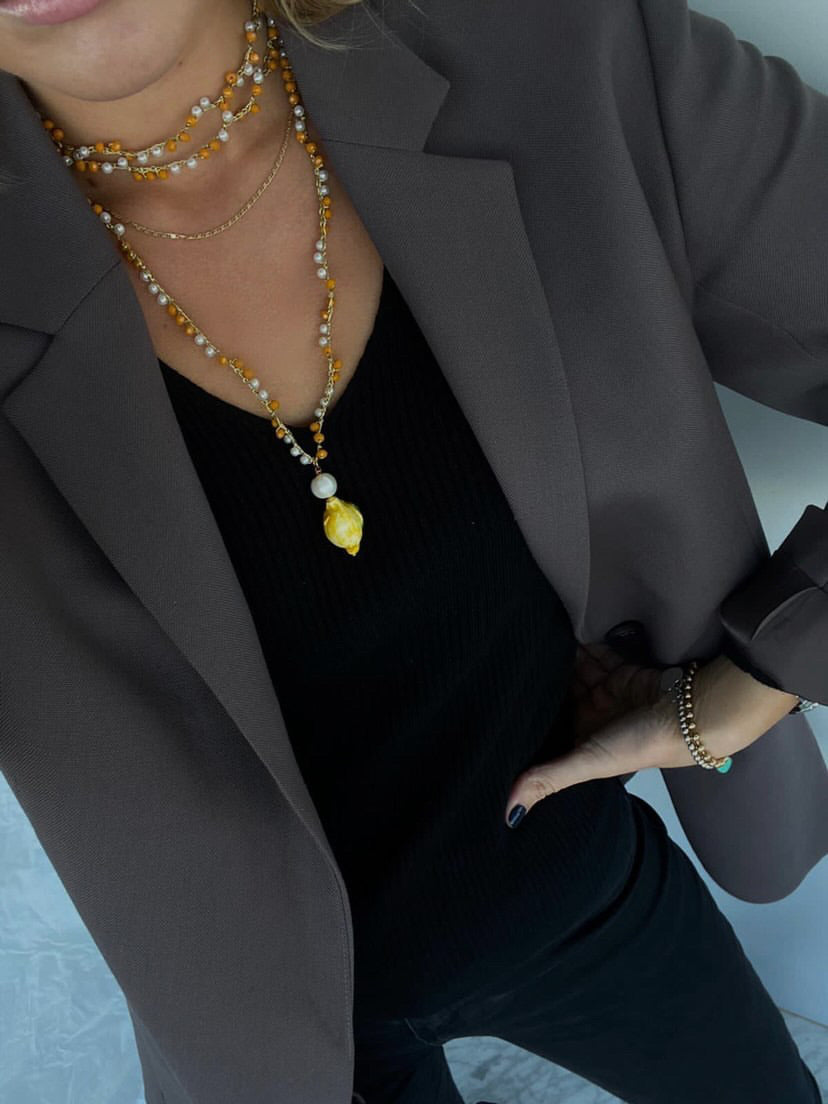 Collar largo con cuentas en tonos cítricos y colgante esmaltado en forma de limón degradado y cierre de perla, con dos vueltas al cuello, combinado con conjunto negro y americana gris. Cashfana.