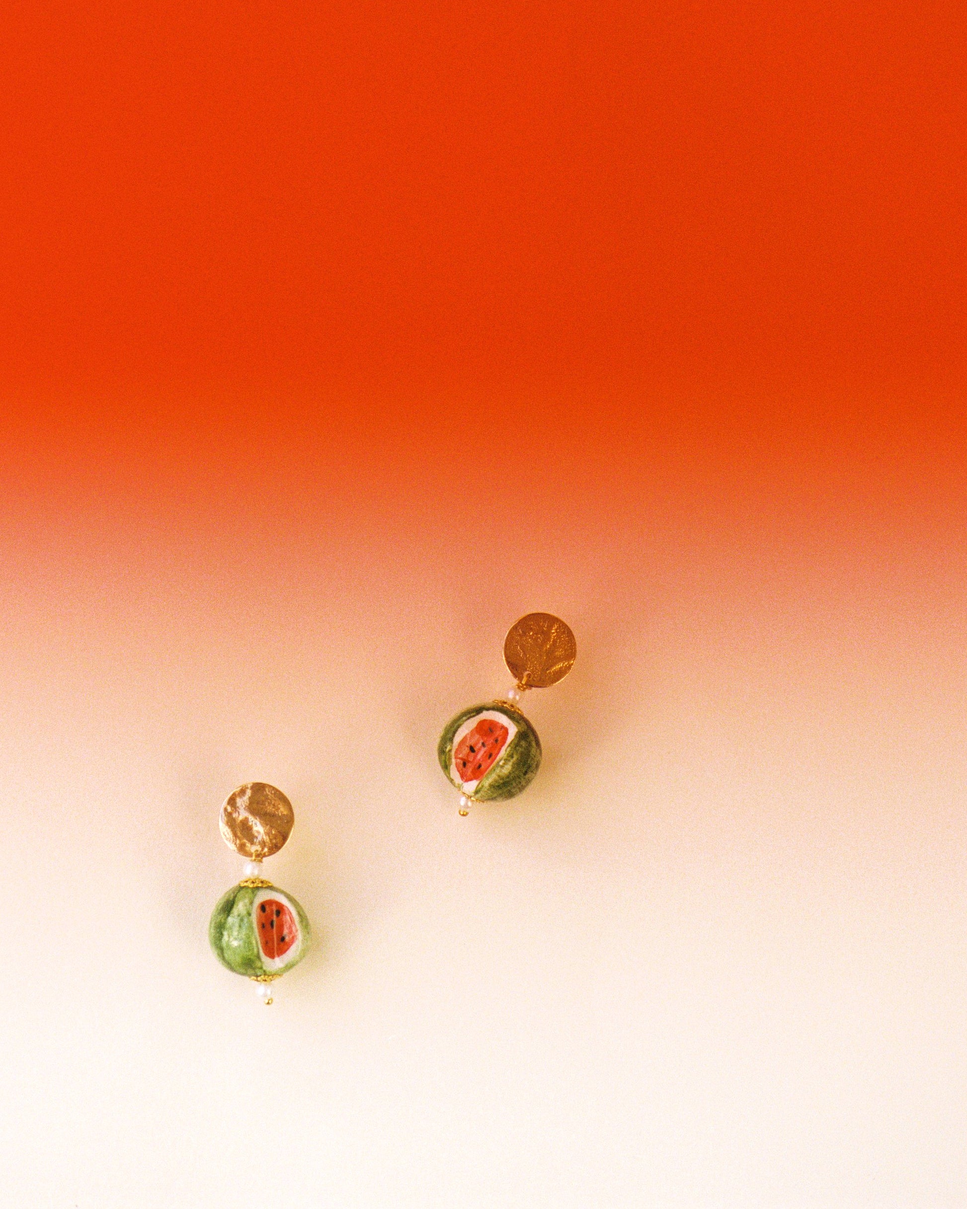 Pendientes con disco dorado y colgante de cerámica con diseño de sandía pintada a mano. Sobre fondo rojo y blanco.

