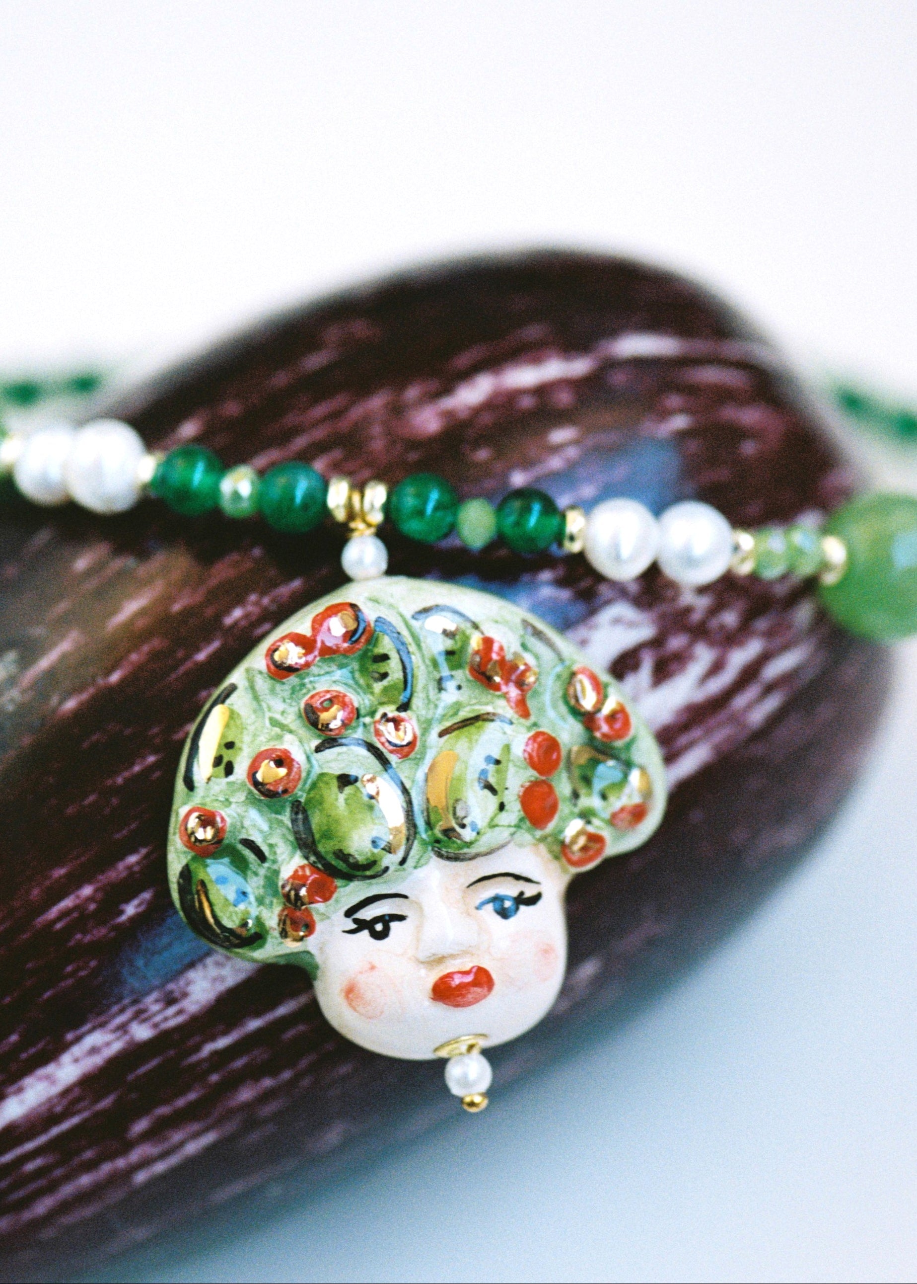 Collar corto con cuentas verdes, perlas  y colgante de cerámica pintado a mano con una carita adornada de hojas y uvas rojas. Cierre dorado con extensión. Sobre berenjena.
