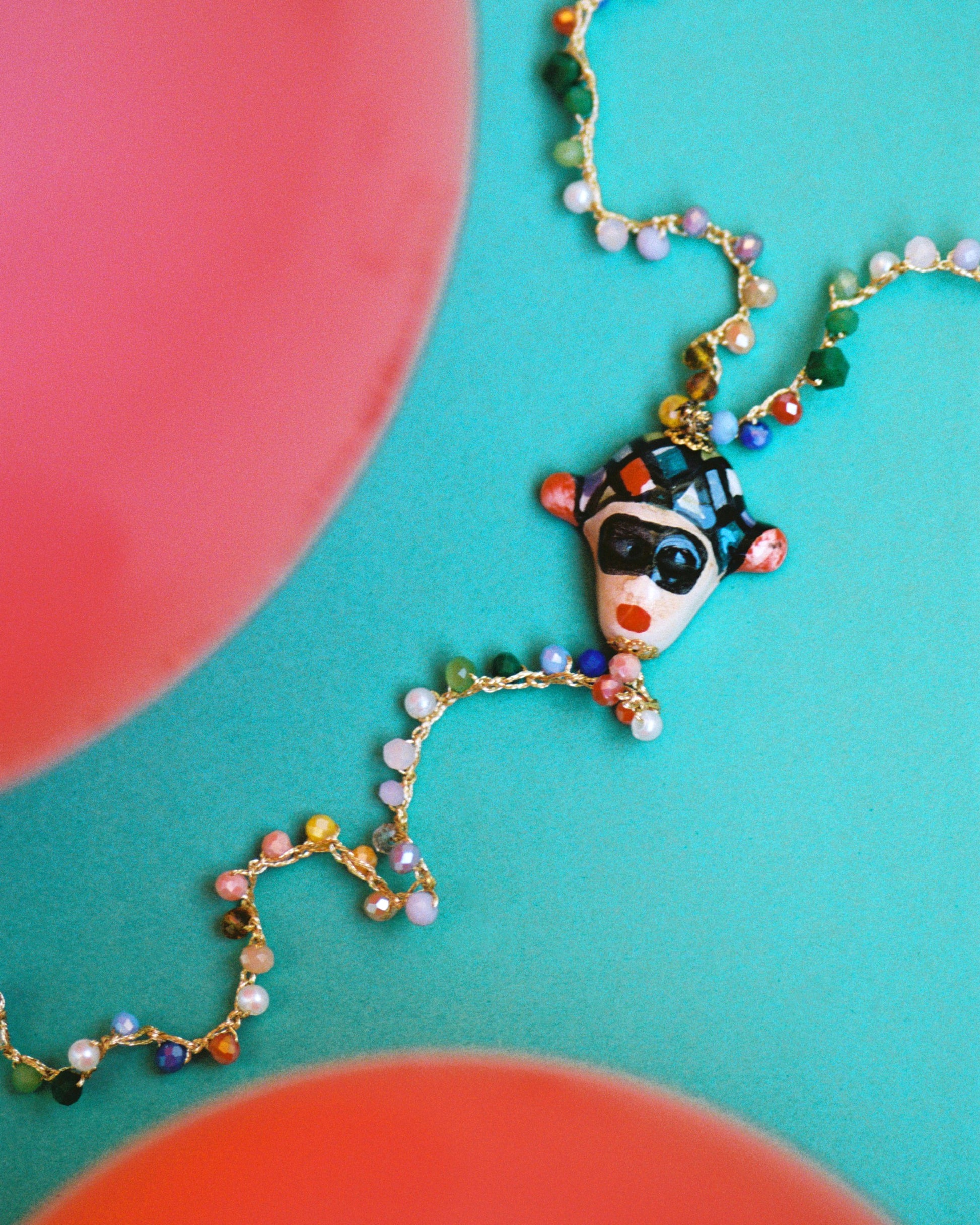 Collar largo multicolor con cuentas de vidrio, figura central de cerámica artesanal con rostro siciliano y dije colgante en forma de cuerno rojo. Sobre fondo azul y rojo.

